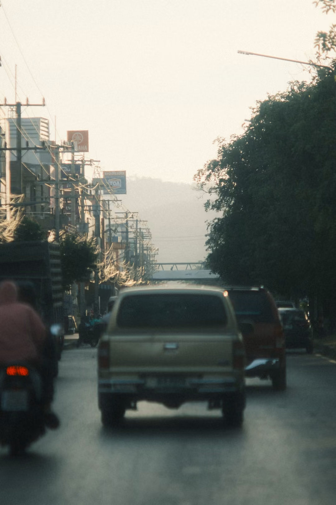 A gold pickup and a motorbike rider in pink head down a hazy town road toward a footbridge and distant hills.