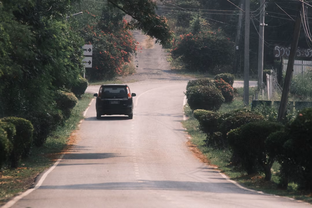 A dark car further along a hedge-lined road climbing toward trees, a road sign and power poles at the sides.