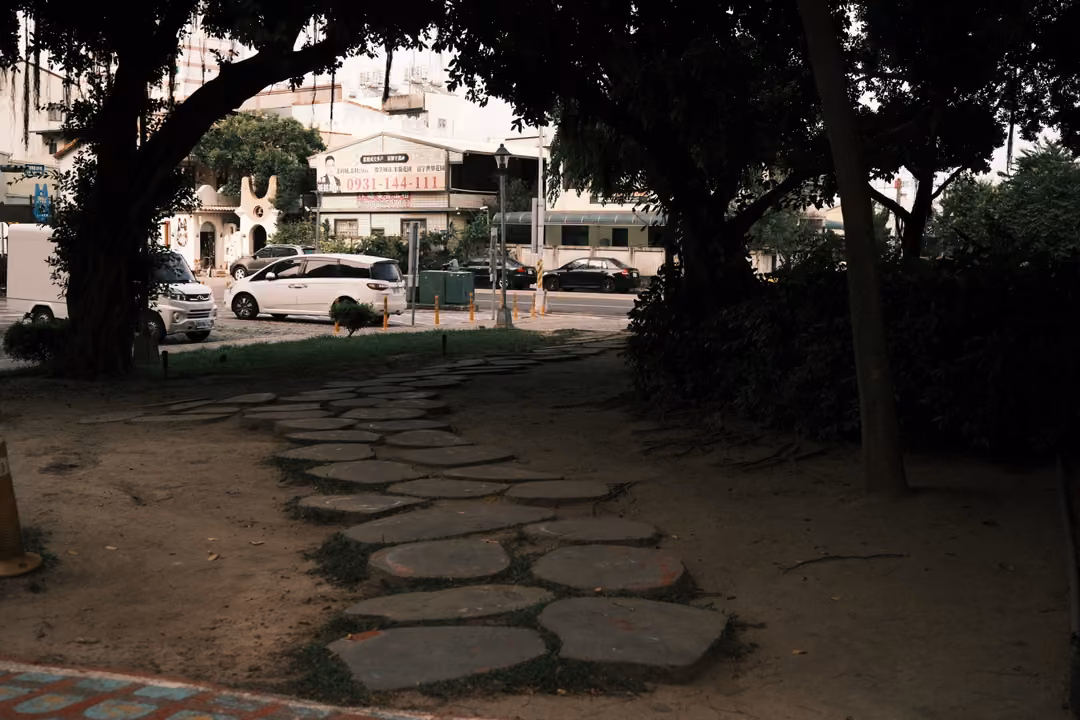 Path of round stepping stones through shaded dirt under trees, a street with parked cars beyond