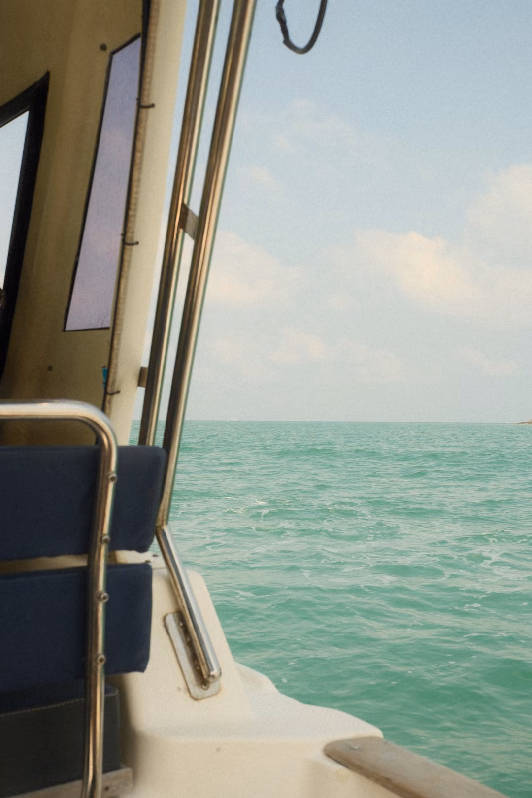 Looking past a boat's cabin, blue seat and chrome rail to turquoise sea and blue sky.