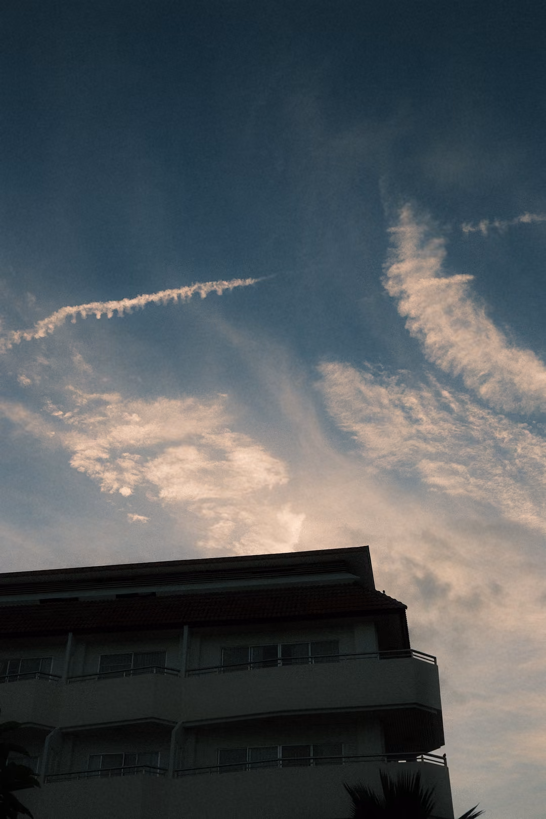 Blue sky with a white contrail and wispy clouds above the dark corner of a multi-storey building