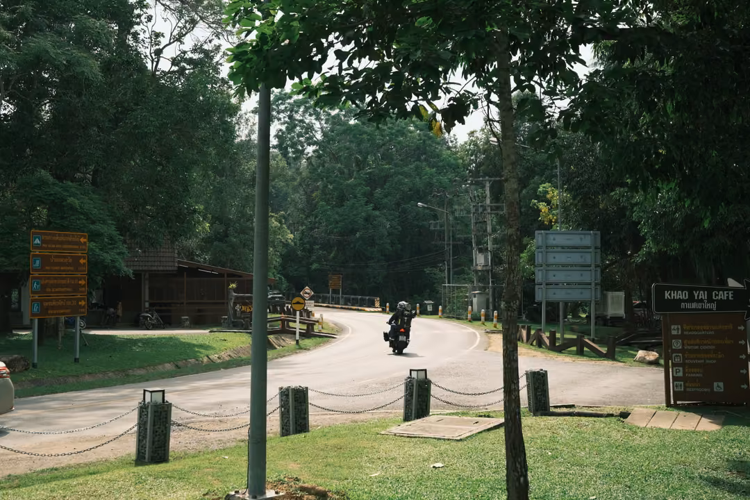 A curving road through trees with a motorcyclist, wooden signposts and a sign reading Khao Yai Cafe
