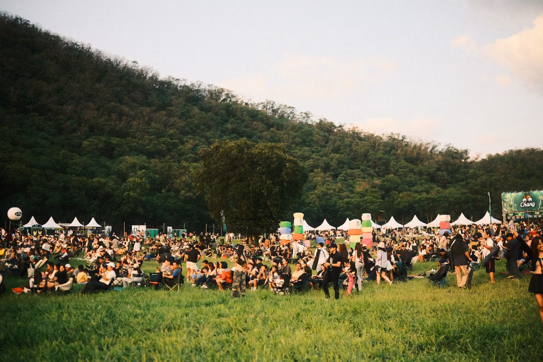 Large crowd seated on a green lawn at the foot of a steep forested hill, white tents behind.