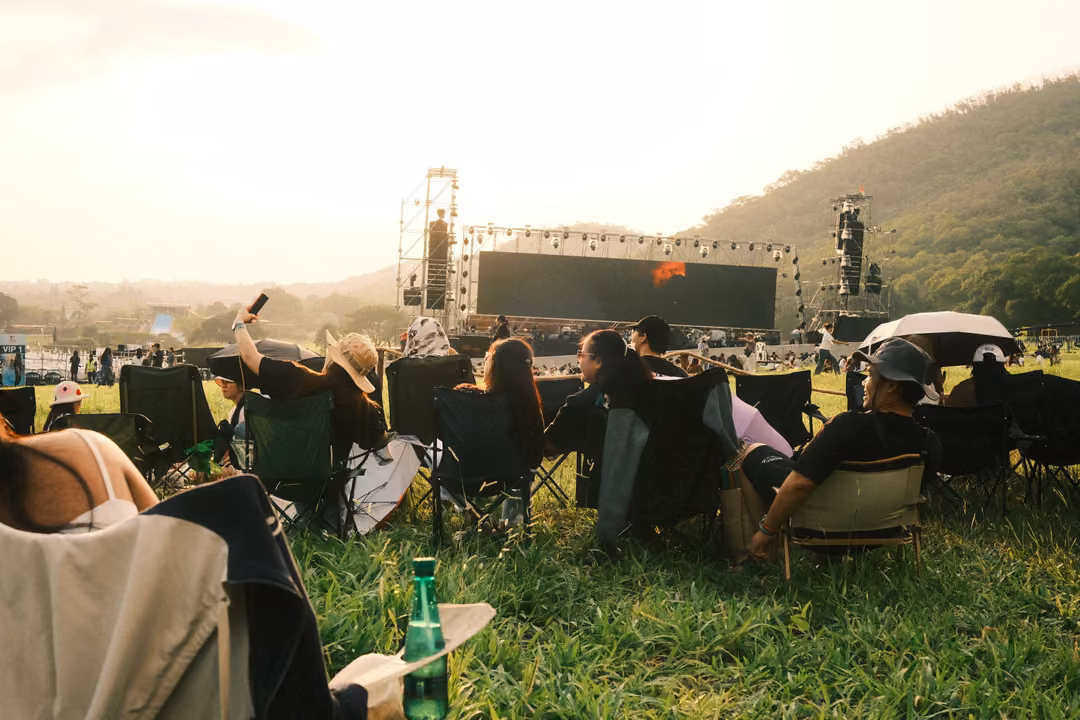 People in camping chairs on a grassy hillside face a large outdoor stage and screen in hazy light.