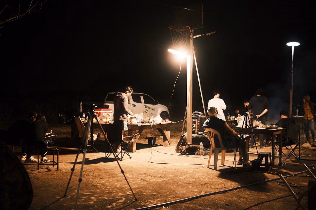 Night campsite with tables, lamps on poles, a tripod, a pickup truck and several people gathered
