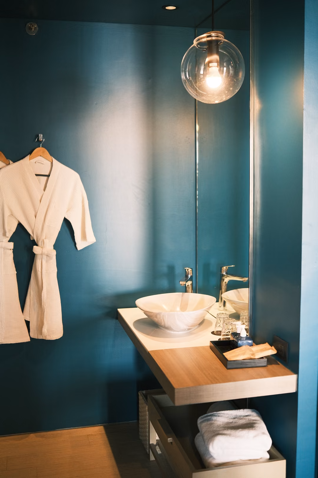 A bathroom alcove with a hanging glass bulb, a white robe and a round basin on a wooden counter