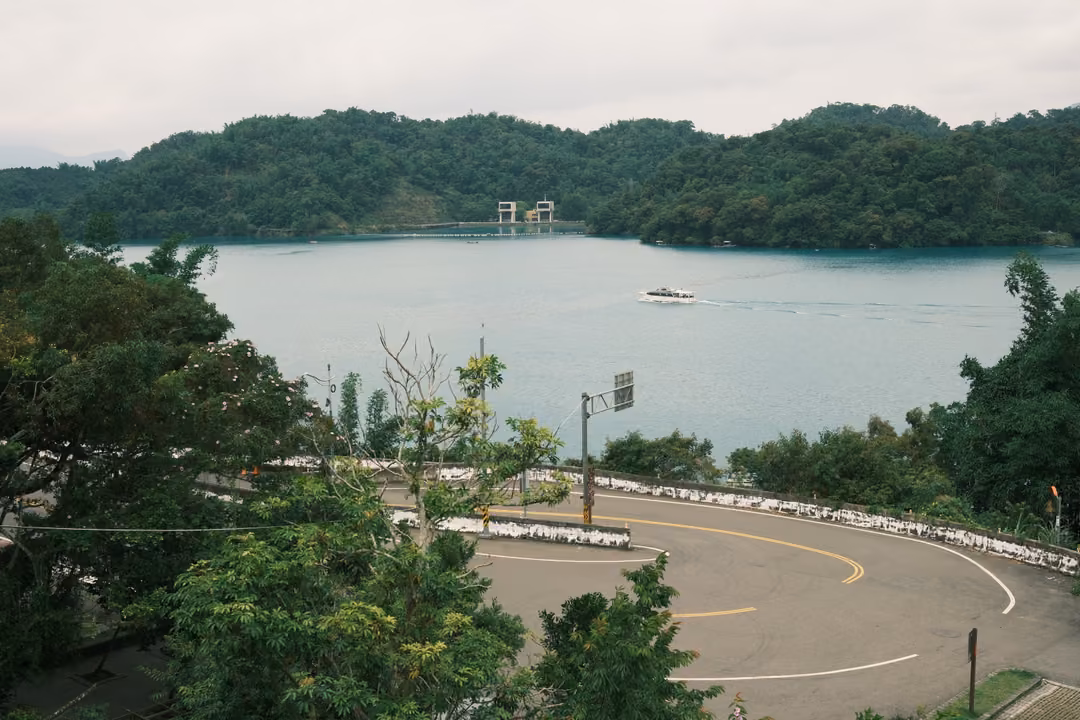 View down to a curving road beside a lake, a white boat crossing toward forested hills