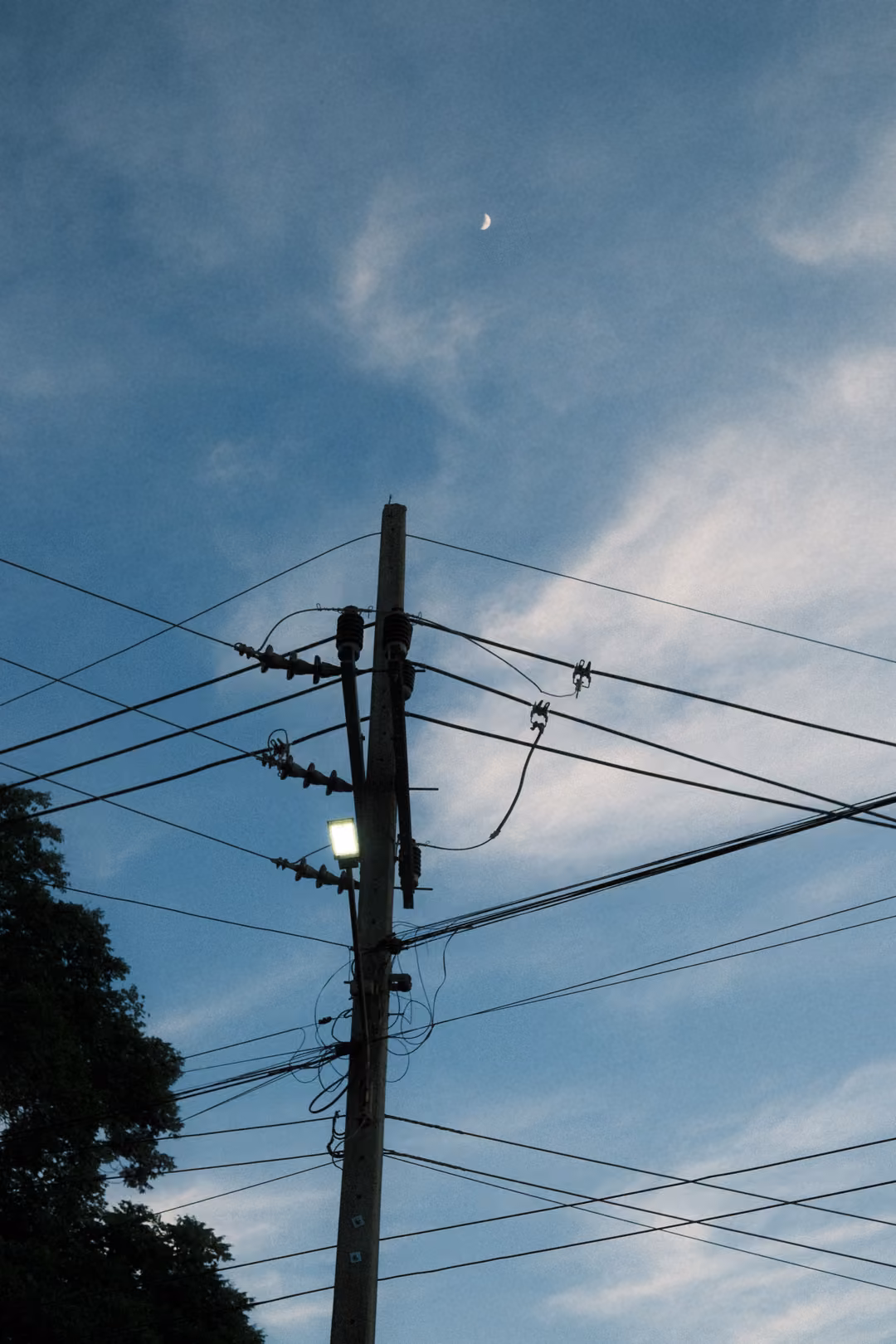 Crescent moon high above a power pole with a lit lamp and tangled wires, dusky blue sky