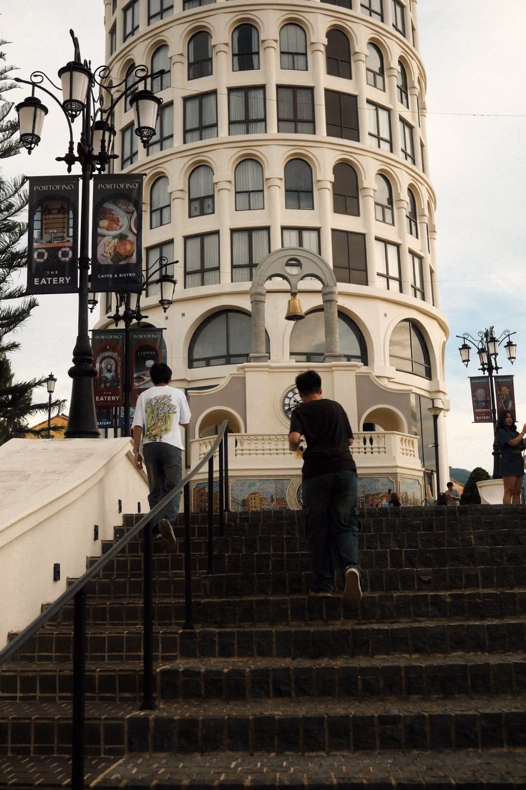 Two people climbing dark stairs toward a tall round tower with arched windows and hanging banners