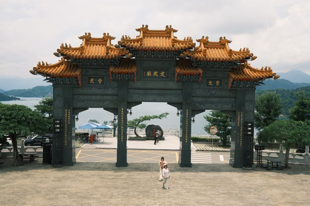 Wider view of the golden-roofed gateway with small figures walking beneath it, lake and mountains behind