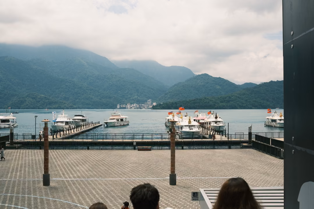 Lake with moored white boats below green mountains under heavy cloud, a paved plaza and heads in the foreground
