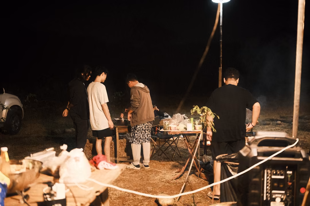 Group of people standing around a table at a night campsite, a lamp post, a speaker and cables on the ground