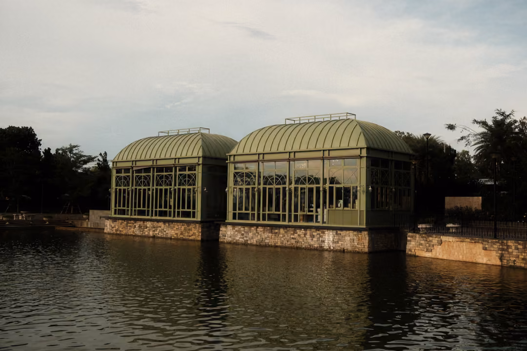 Two glass pavilions with curved green roofs on a stone embankment beside a calm lake under a grey sky