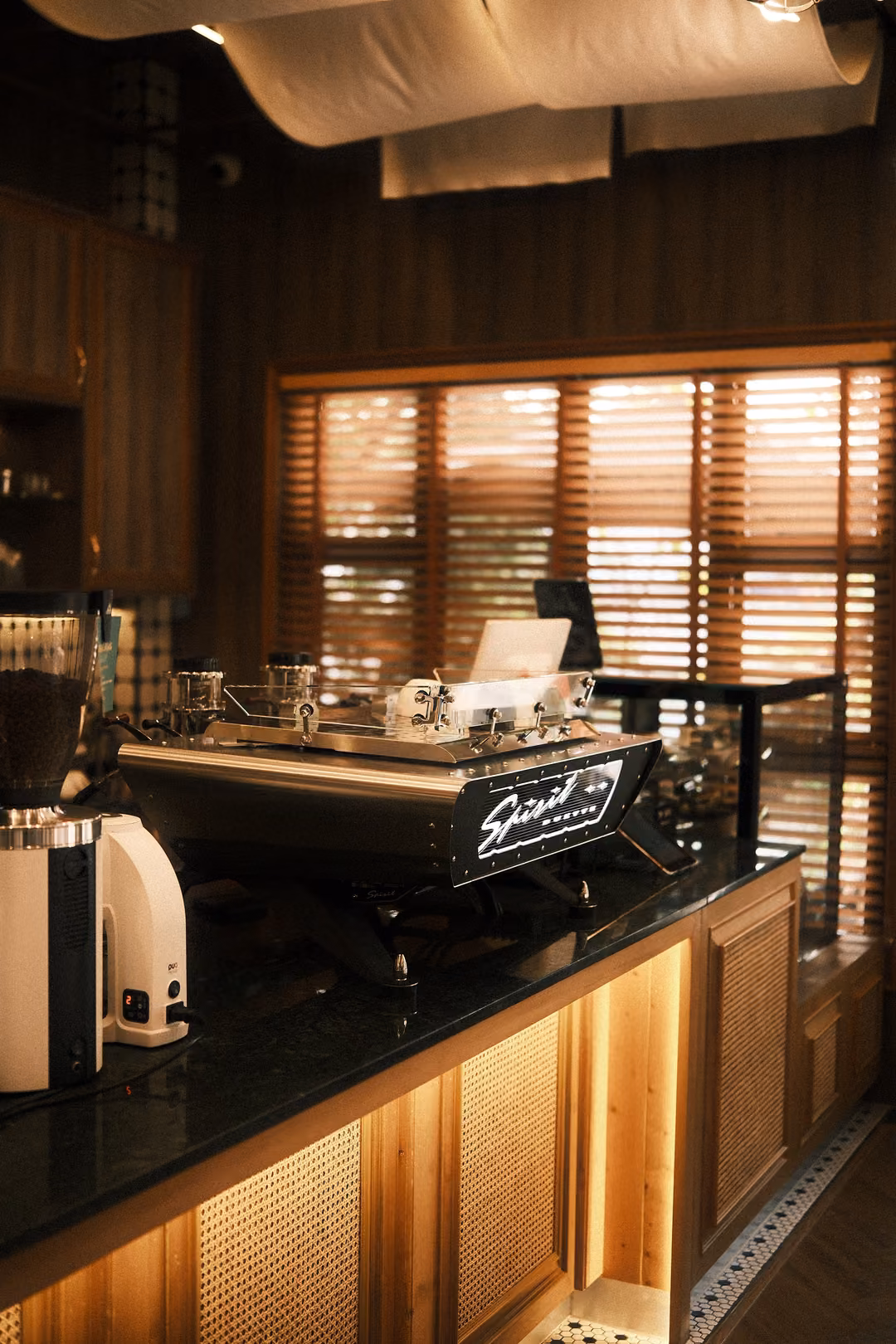 Espresso machine and white grinder on a dark counter under draped fabric, wooden blinds behind