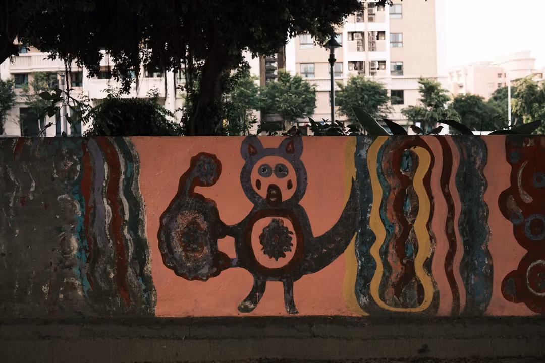 Pink wall painted with a cartoon creature and wavy coloured stripes, apartment towers behind trees