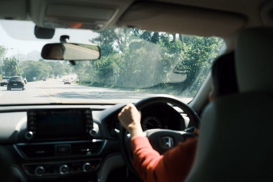 Driver's hand on the steering wheel, multi-lane road with cars and roadside trees through the windscreen