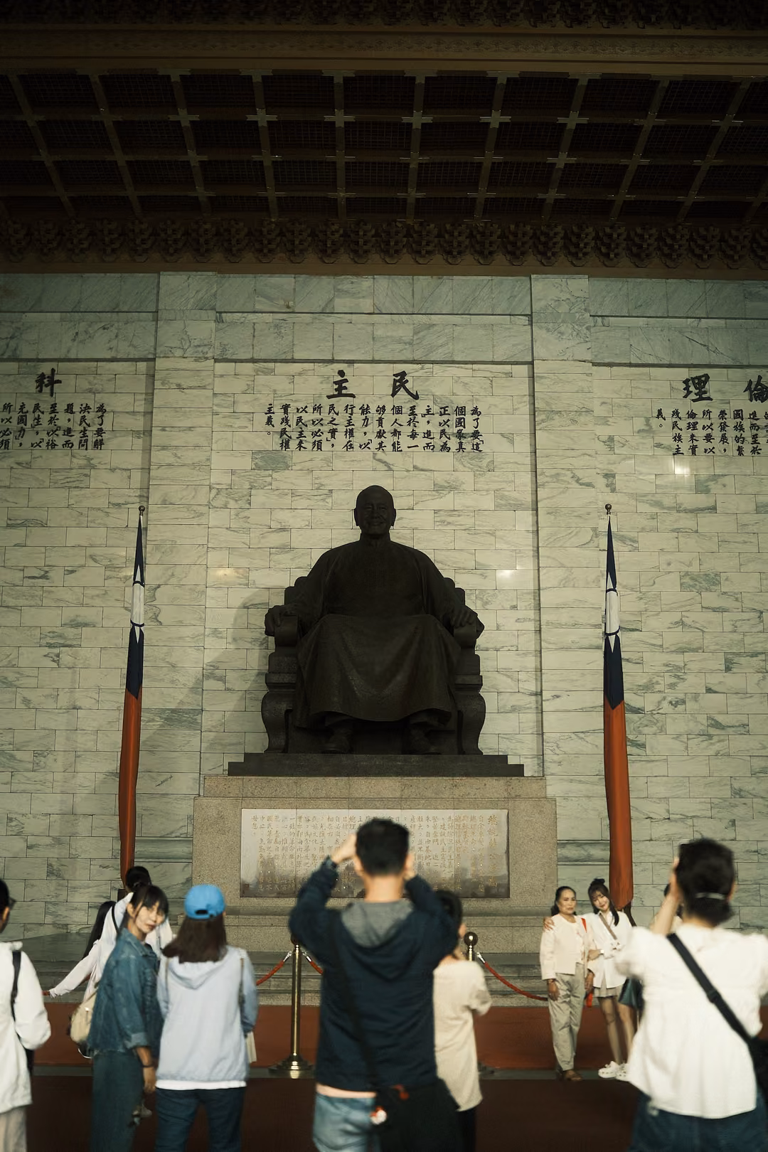 Visitors with phones raised before a seated bronze statue flanked by two flags in a marble hall