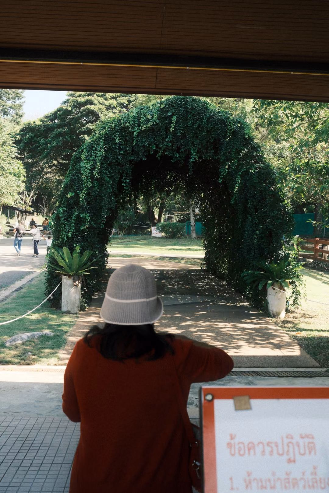 Person in a beige bucket hat and red top looking at a leafy arch, a Thai rules sign in the corner