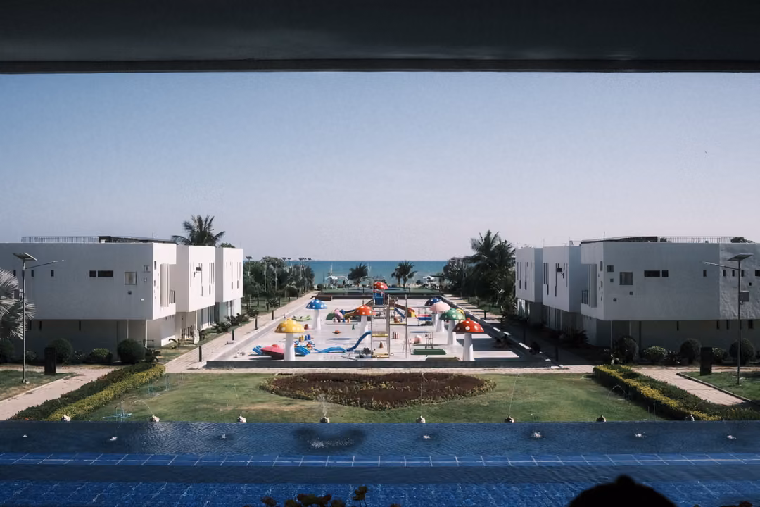 View from a covered balcony over a blue pool to white buildings, a colourful splash park and the sea beyond