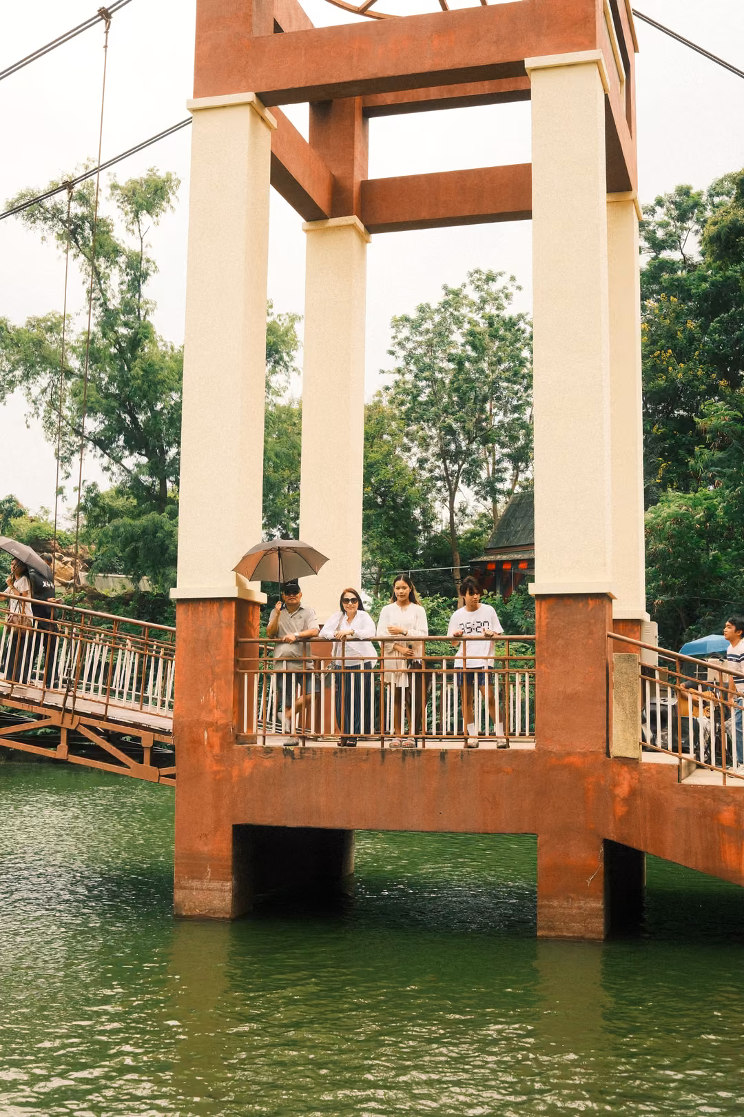 Red and white bridge tower rising from a lake, people with umbrellas standing on the walkway between its columns