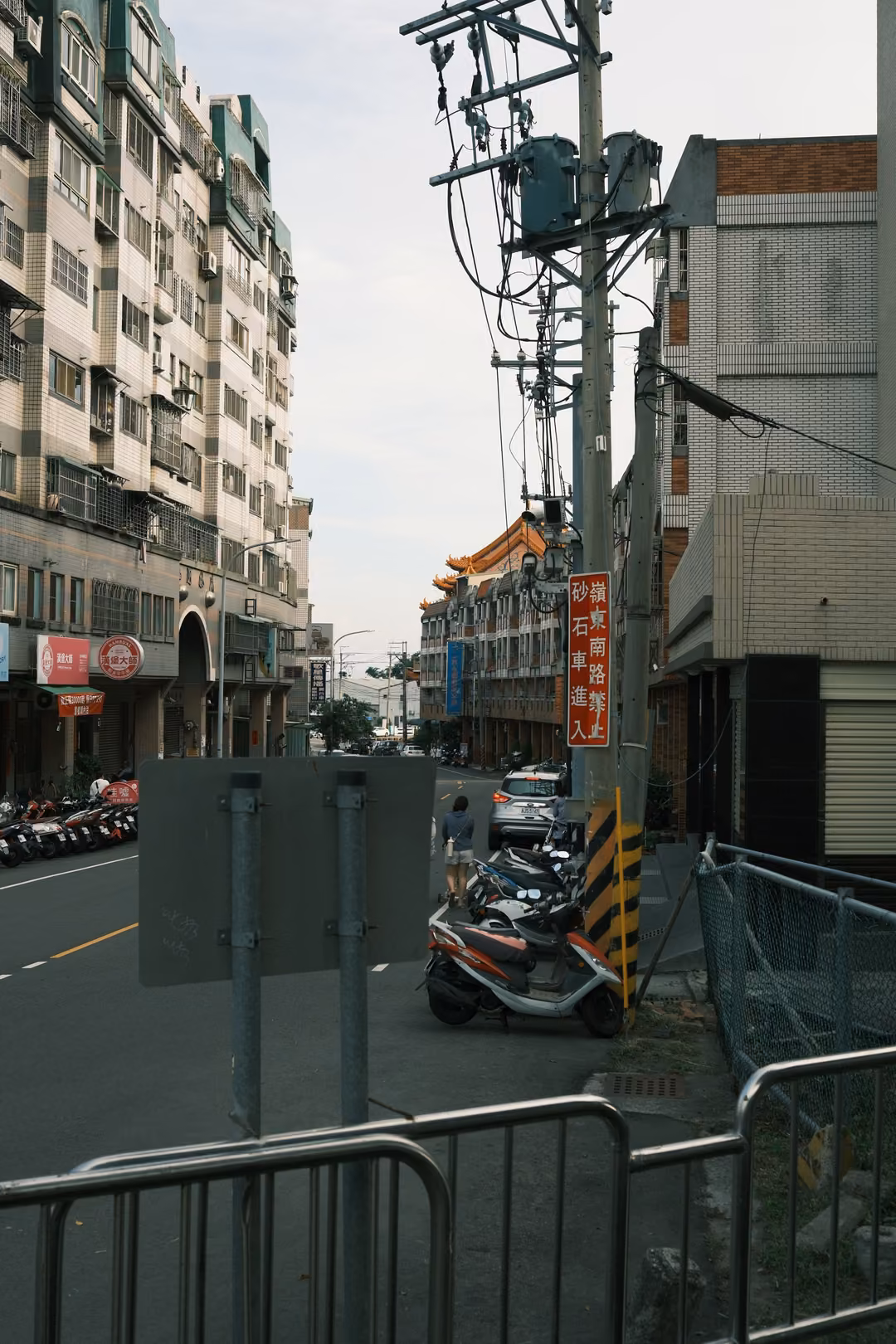 Side street with tall buildings, a utility pole with transformers, metal barriers and a parked scooter