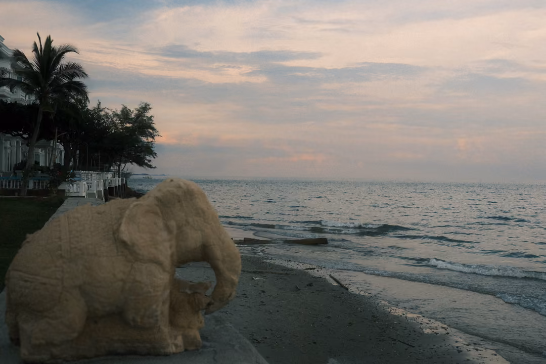 Stone elephant sculpture in the foreground on a seawall, waves and palm trees along the shore under evening clouds