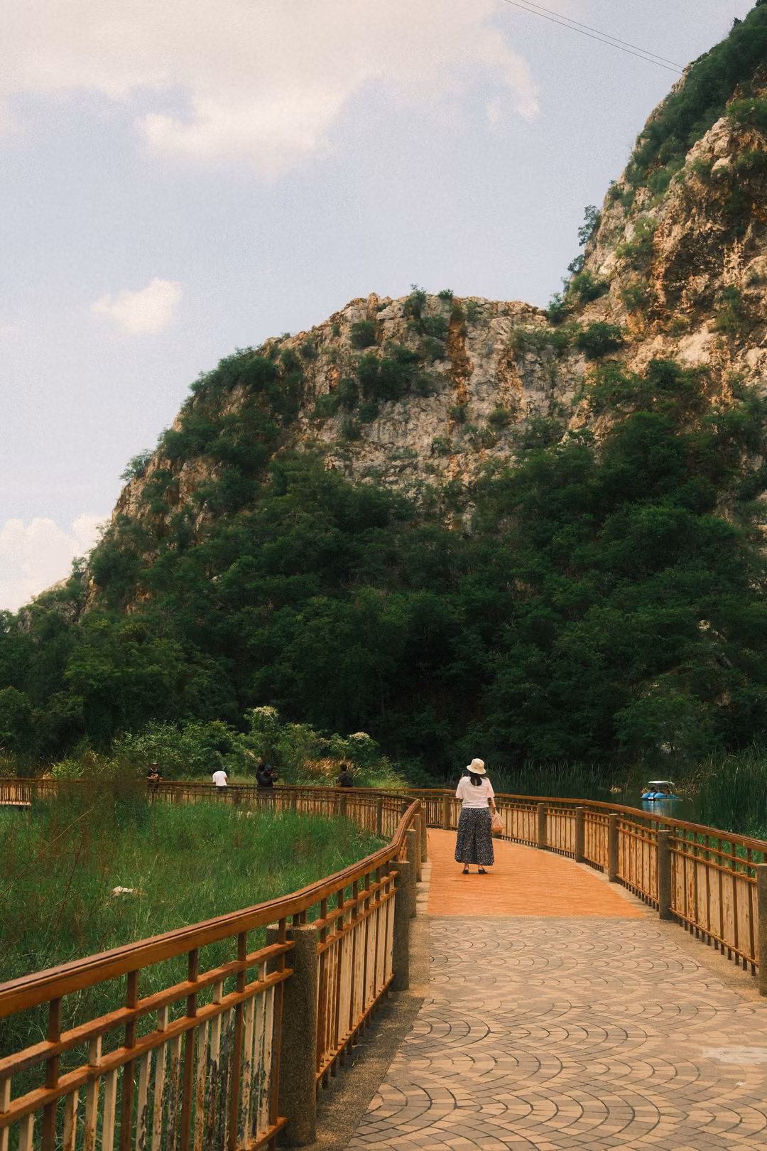 Curving paved walkway with wooden railings, a woman in a sun hat walking ahead, green hill under blue sky