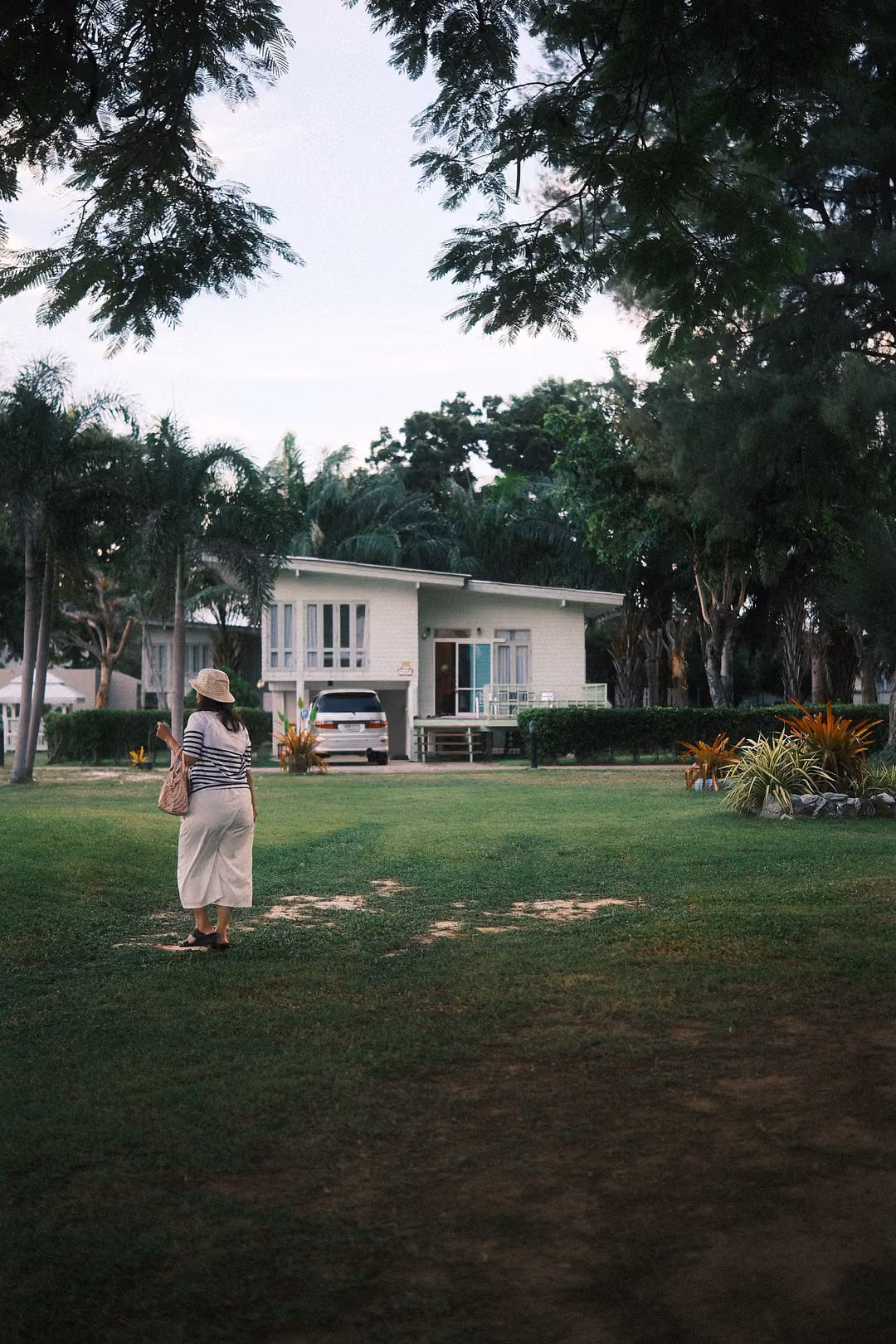 Woman in a sun hat and long white skirt standing on a lawn facing a white house with a parked car