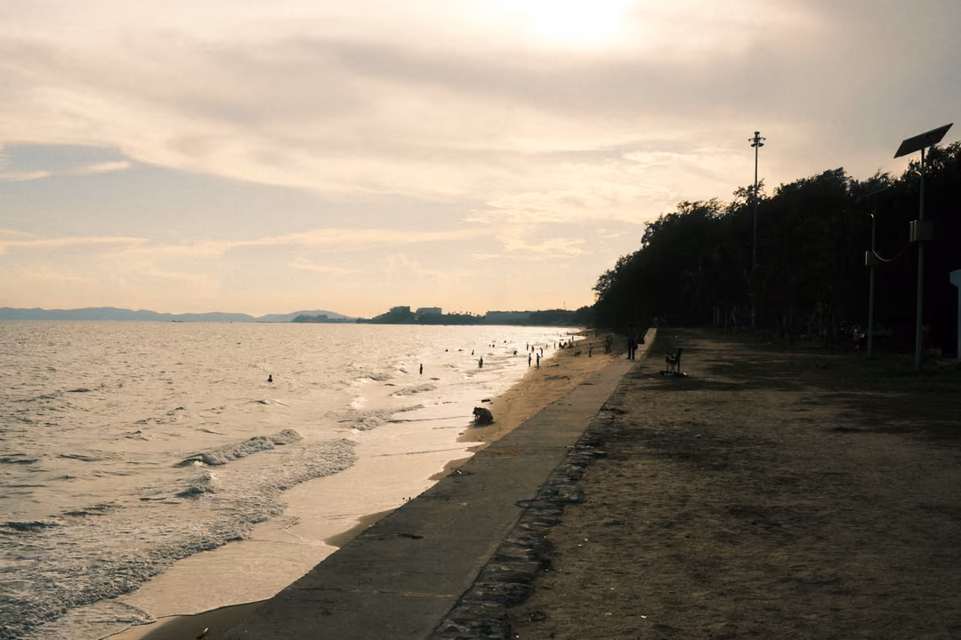 Beach and seawall at dusk under a hazy glowing sky, trees and lamp posts along the shore, swimmers far off