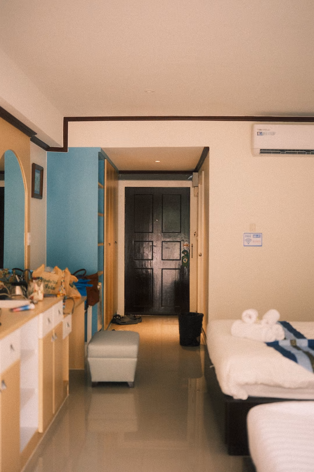 Vertical view of a hotel room hallway with beds, a cabinet, a white ottoman and a dark wooden door