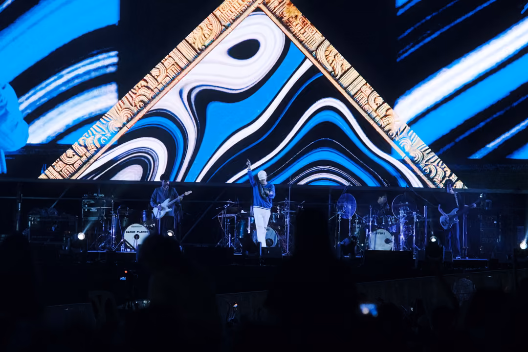 Singer in a blue jacket and white trousers raising an arm below a gold-framed triangle of swirling blue stripes.