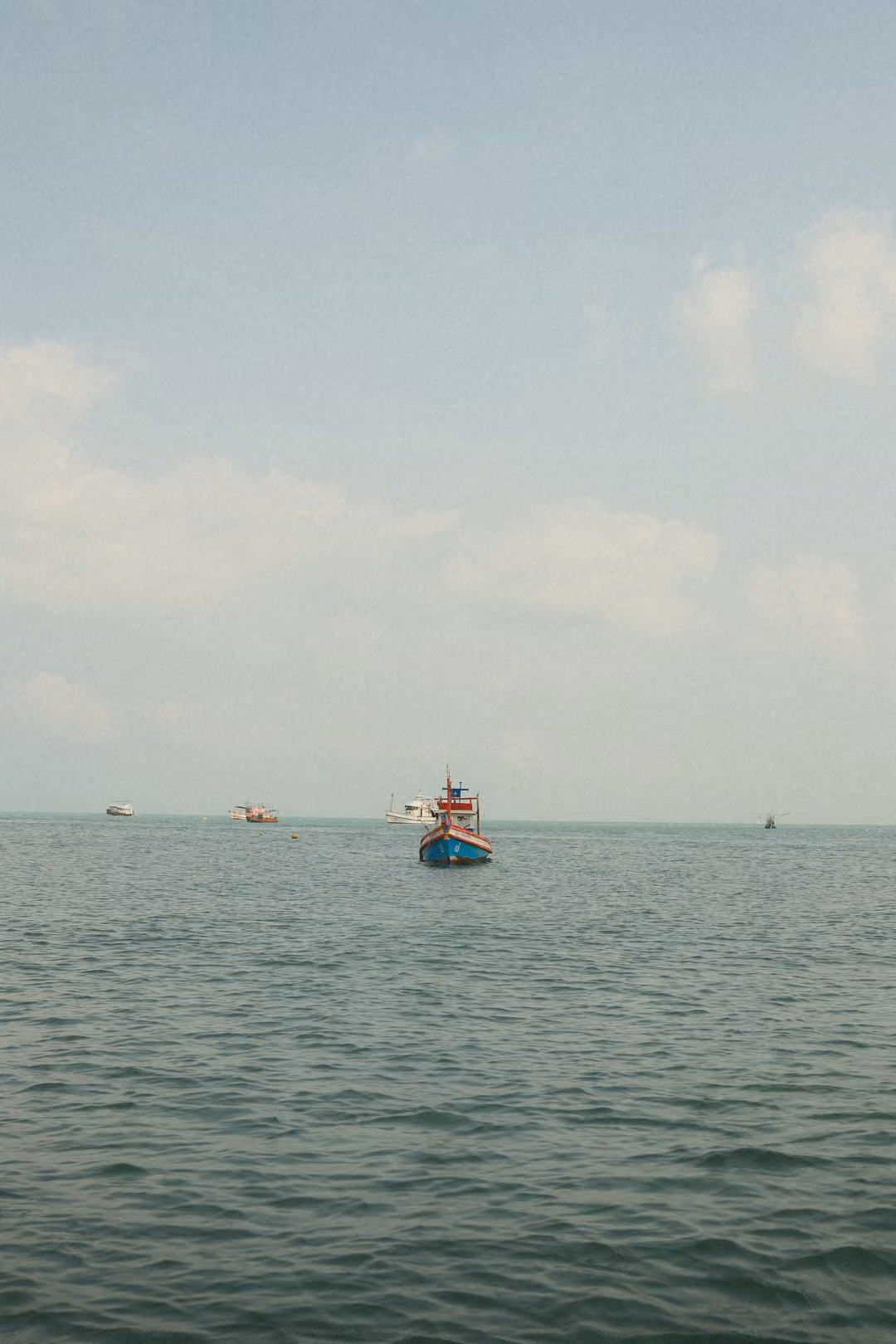 A small painted fishing boat alone on open grey-green sea, with distant boats on the horizon.