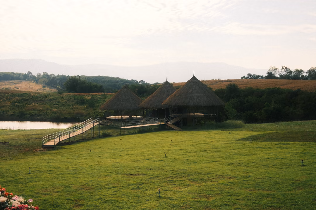 Thatched-roof huts on stilts beside a pond, linked by a wooden ramp, with green lawn and hills behind.