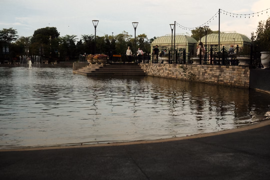 Wide pond with a small fountain, stone steps, lamp posts, string lights and green-roofed pavilions beyond