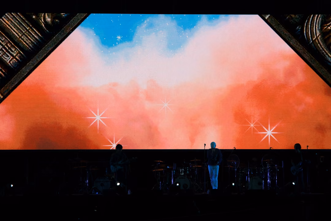 Wide screen of orange and pink clouds with sparkles, gold carved corners, the singer standing small on stage below.