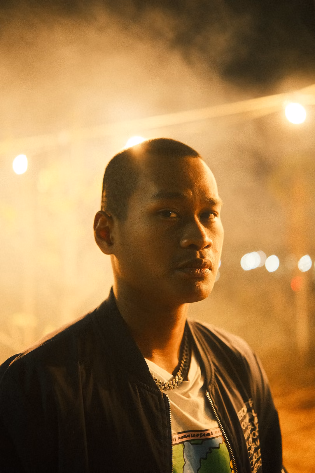 Vertical portrait of a person in a dark jacket and chain necklace, smoky golden haze and lights behind