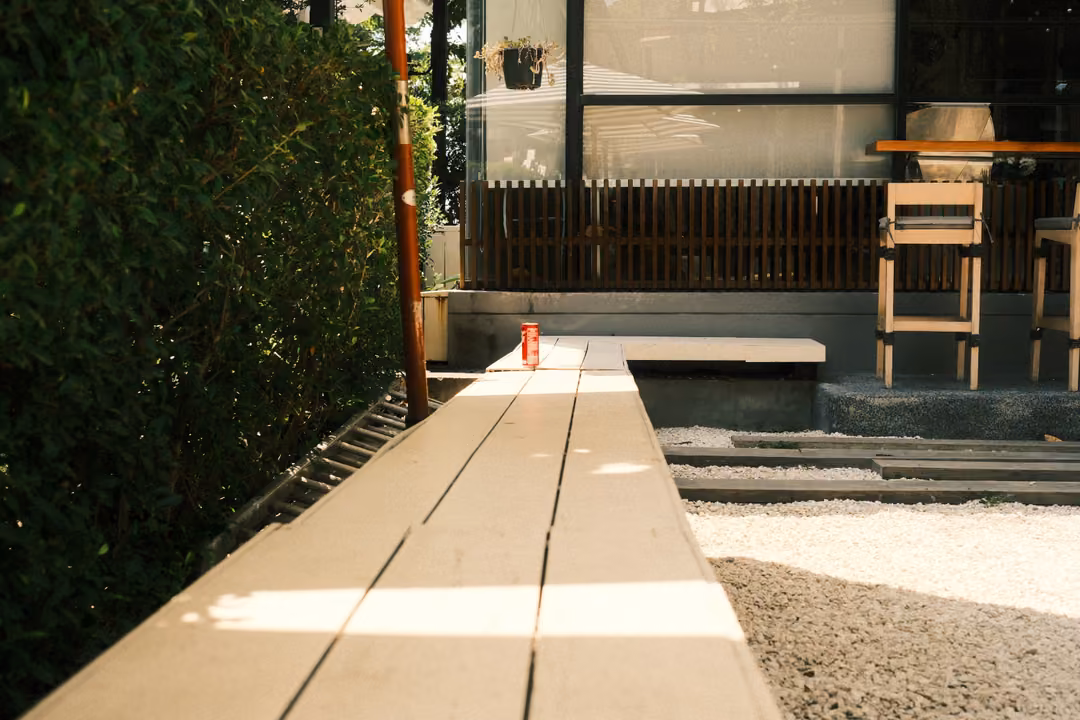 Wooden boardwalk leading toward a glass-fronted building with a bench and stools, hedge on the left, gravel right