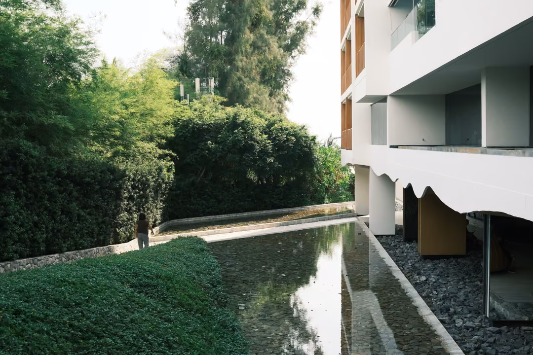 A long reflecting pool between a white building and a dark green hedge under a pale sky