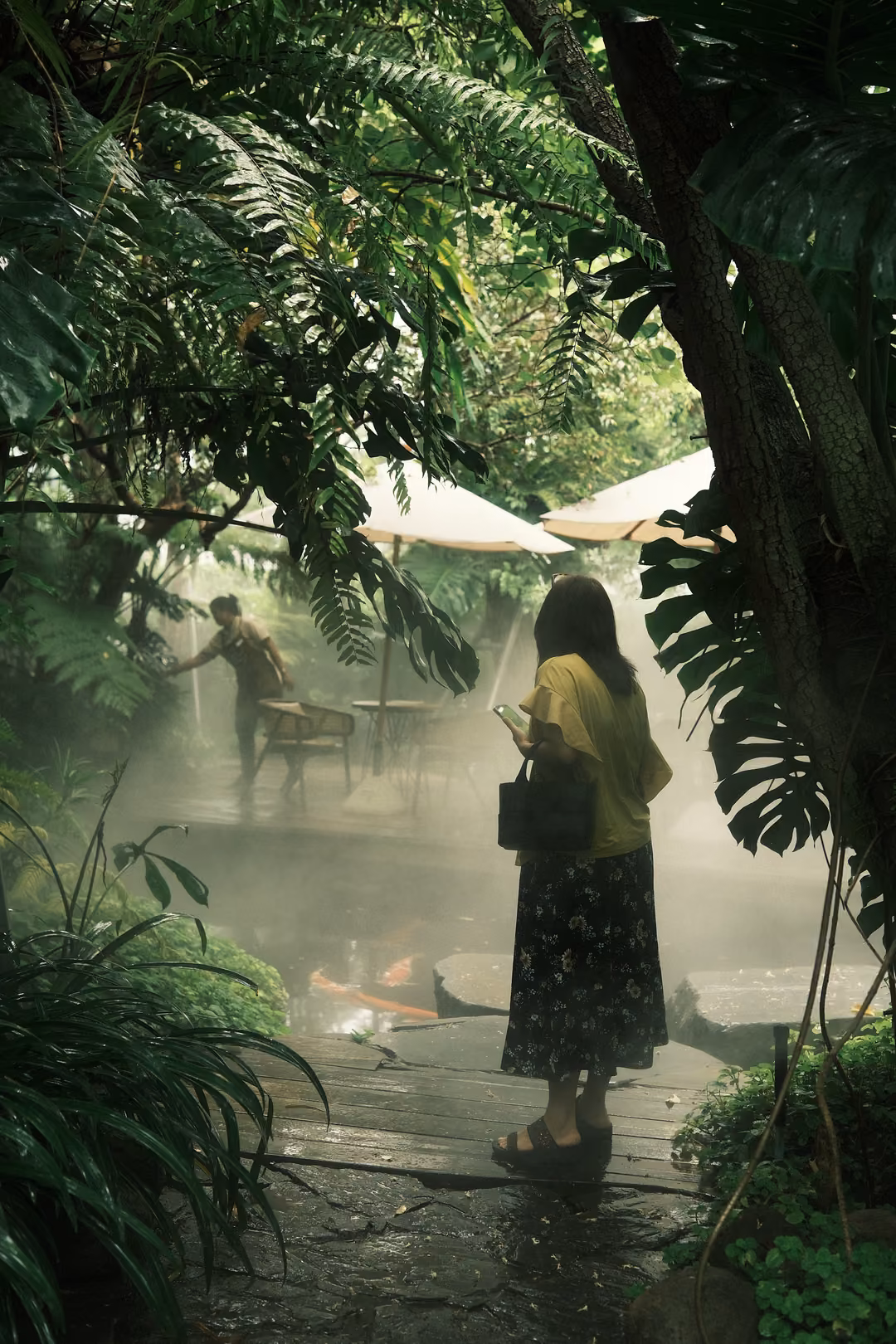 A person in a yellow top holds a phone near a misty koi pond, ferns and umbrellas in the background.