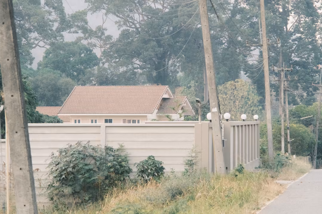 Pink-roofed house behind a tall concrete wall with round lamps, power poles and leafy trees