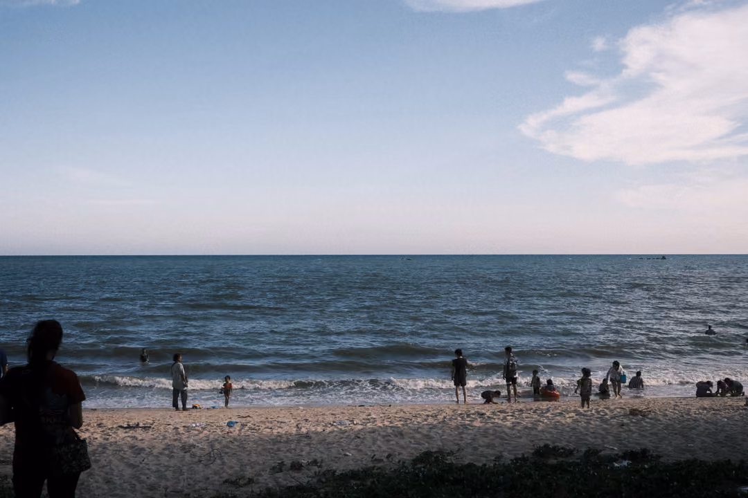Beach with people wading and playing in gentle waves, a figure silhouetted in the left foreground
