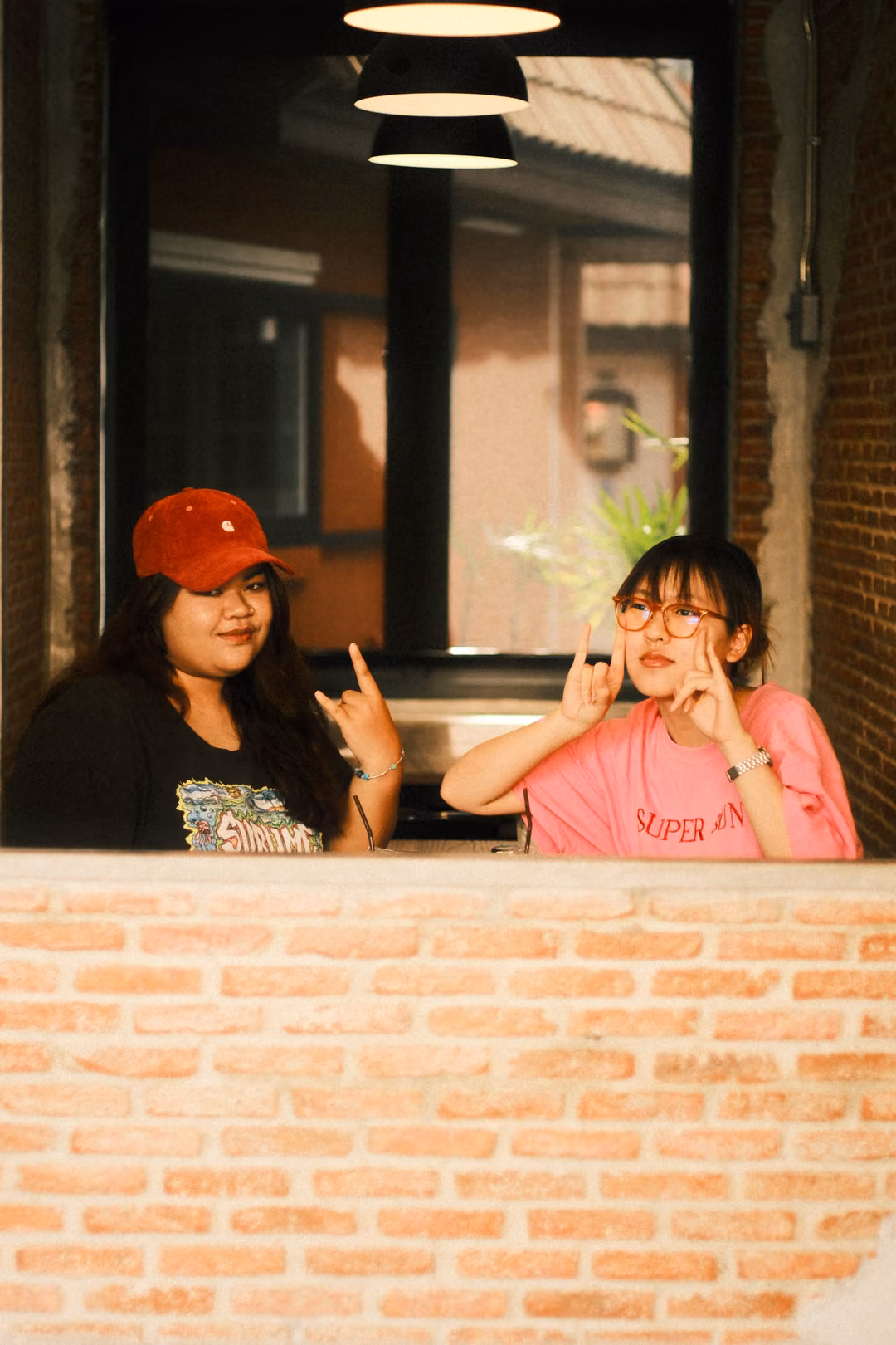Two women behind a brick wall at a window, one in a red cap making a rock sign, the other in pink with glasses posing