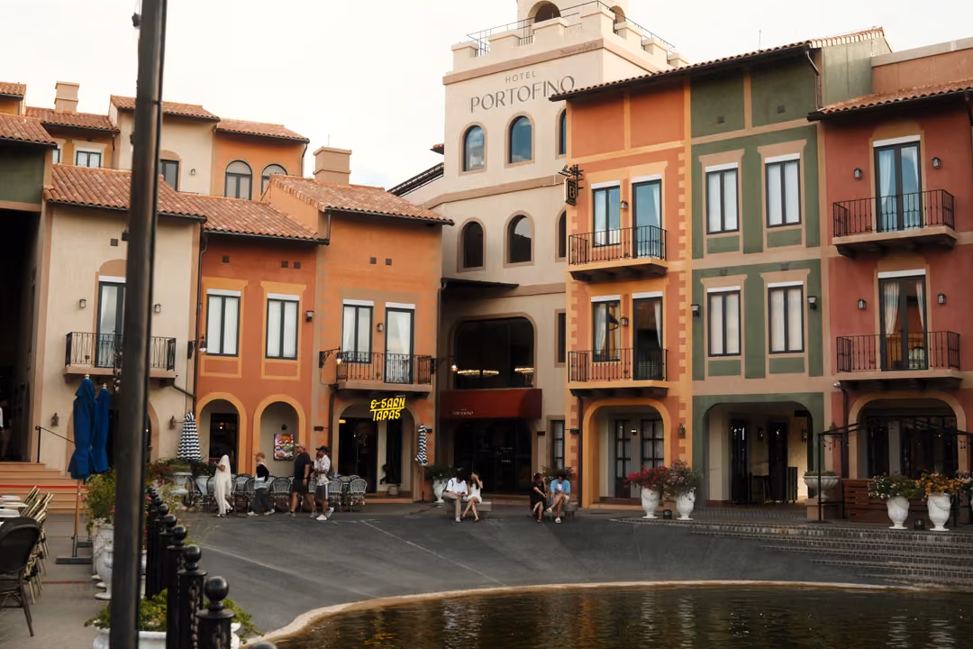 Row of colourful facades including a cream building marked Portofino, with people seated near a pond edge