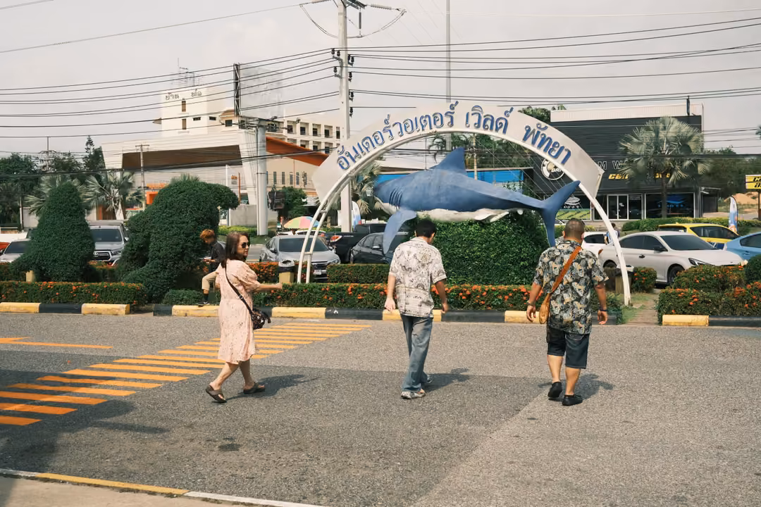 Three people walking across a car park toward an arch with Thai lettering and a blue fish statue