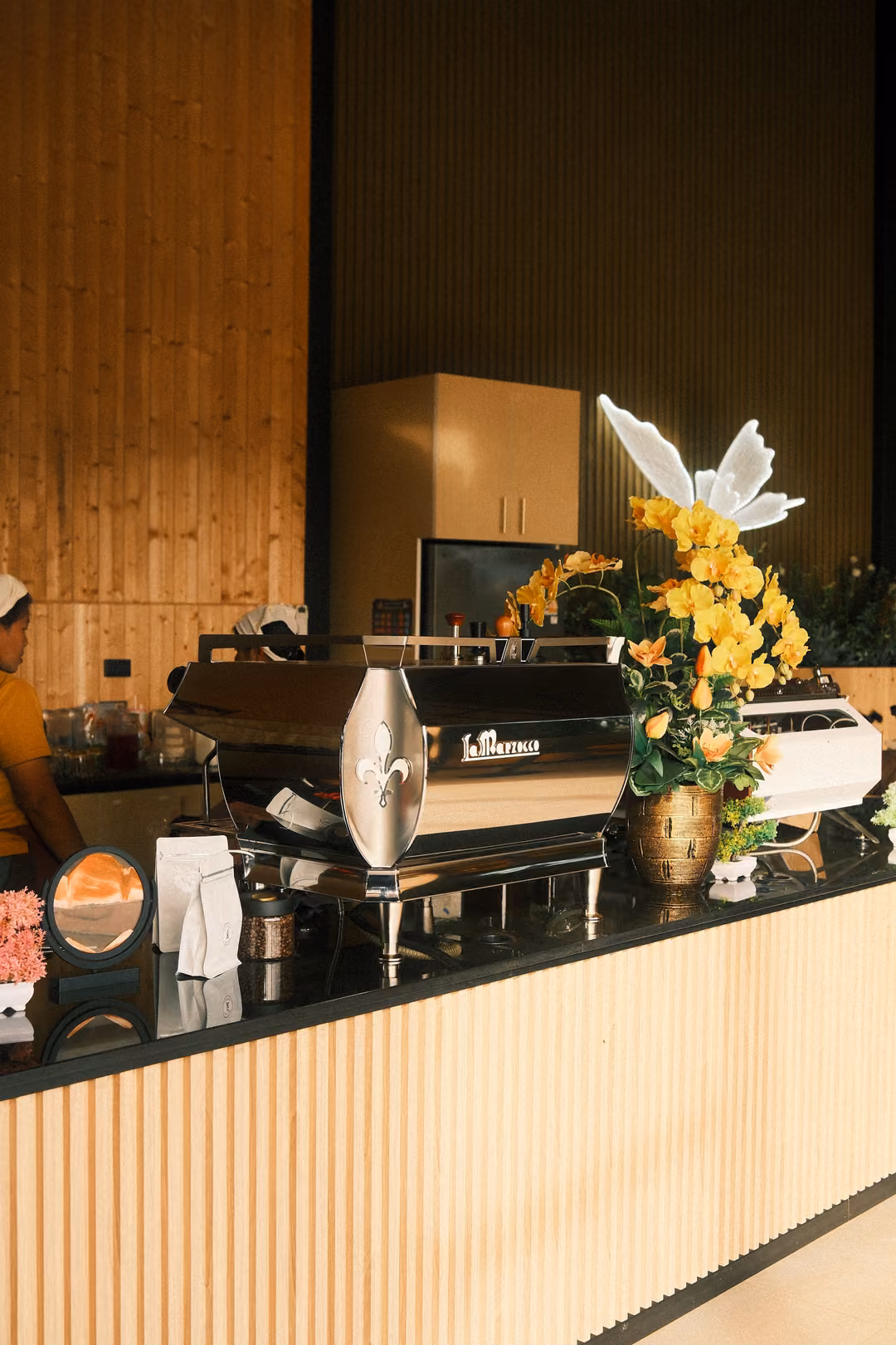 A café counter with an espresso machine, a vase of yellow flowers and a white butterfly ornament.