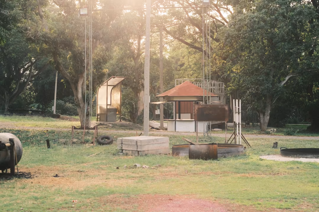 A grassy yard with a gas tank, concrete blocks, a metal pit and a small red-roofed hut, sunlight flaring through trees.