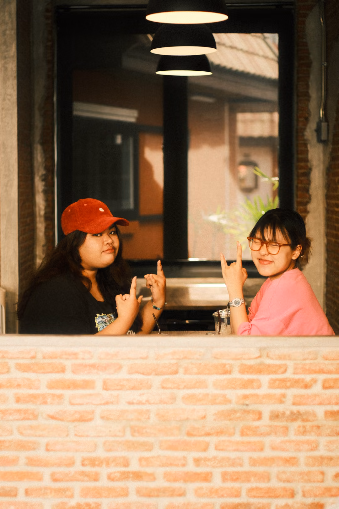 Two women posing at a window, the one in pink making a horns hand sign and grinning, the other in a red cap smiling