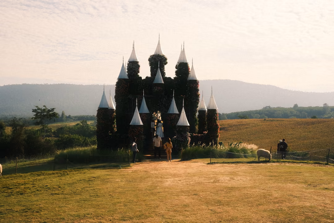 A wide view of the flower-covered castle with white spires, visitors at its base and a sheep to the right.