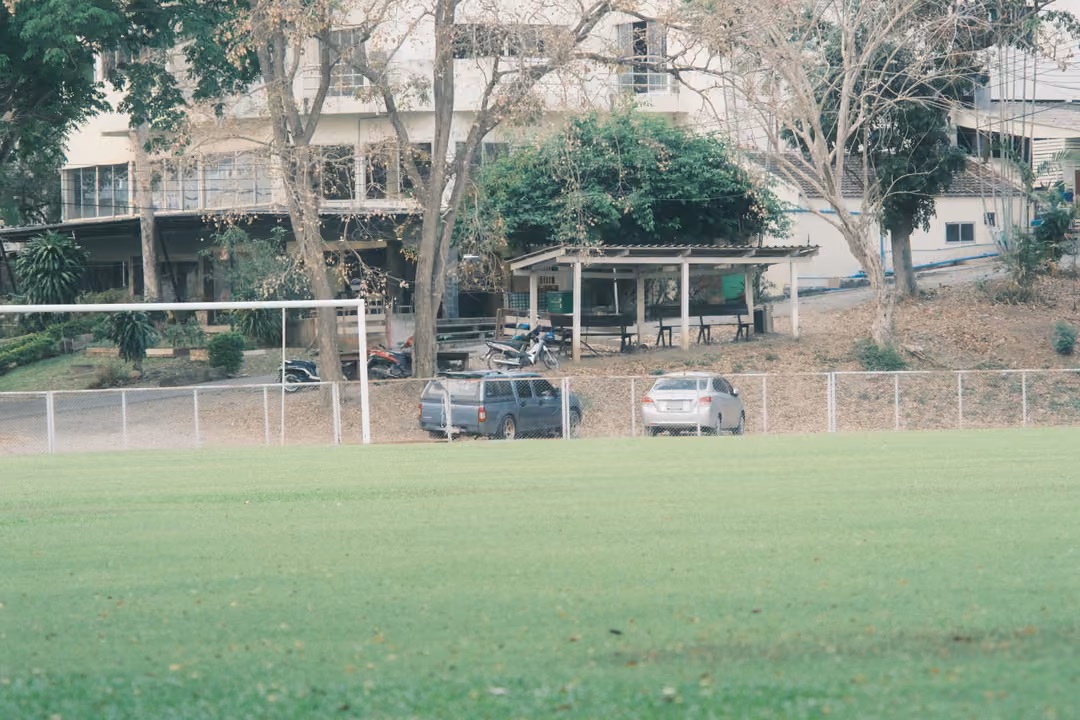 Green sports field with a white goal frame, cars parked by a fence and a pale building among trees