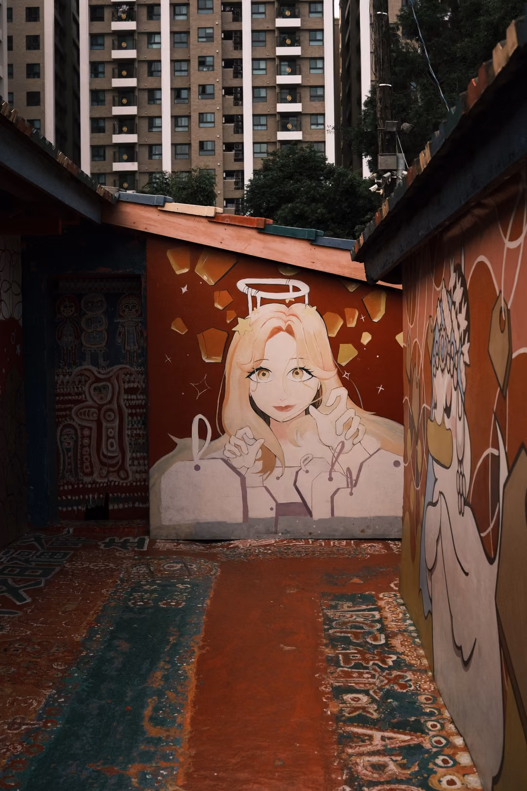 Narrow painted alley with an anime-style angel portrait on a red wall, high-rise apartments rising behind